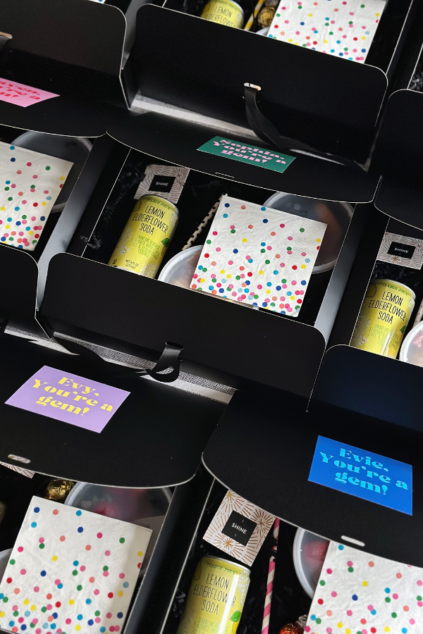 A group of cute black boxed lunches neatly arranged, each with colorful polka dot napkins, a can of lemon elderflower soda, a "shine" fortune card, and custom stickers on the lids with the phrase "You're a Gem!" all tied together with a black ribbon.