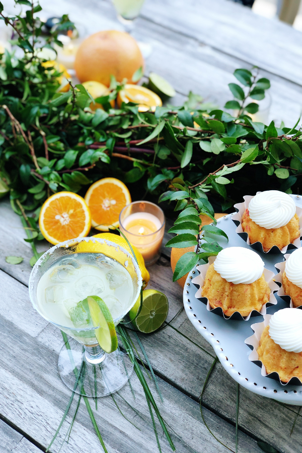 A citrus tablescape with fresh greenery, sliced citrus fruits, and glowing votive candles are artfully arranged along the wooden table with an inviting margarita by it's side signaling a citrus bachelorette theme party.