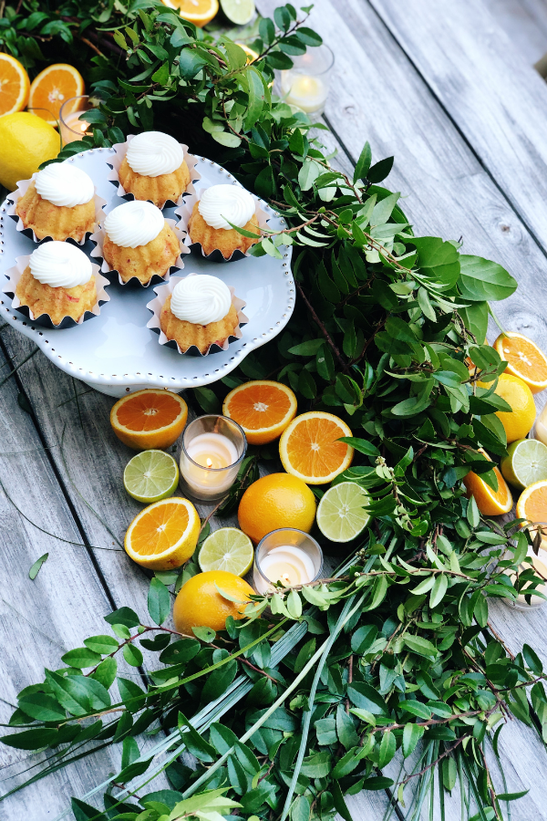 A citrus-inspired tablescape with a white pedestal cake stand showcasing mini Bundt cakes topped with white frosting. Fresh greenery, sliced lemons, limes, and oranges, and glowing votive candles are artfully arranged along the wooden table for an inviting and elegant display as beautiful citrus table decor.