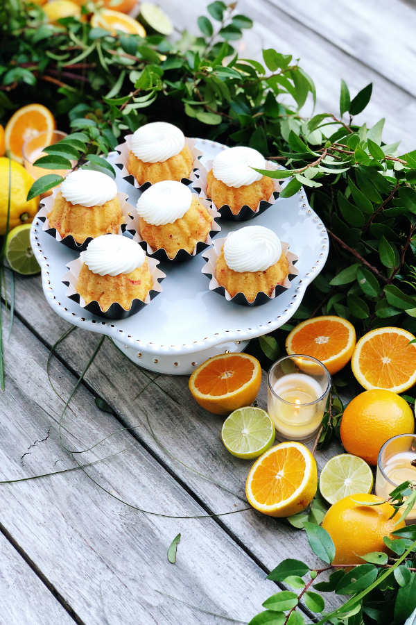 A citrus tablescape with a white pedestal cake stand showcasing mini Bundt cakes topped with white frosting. Fresh greenery, sliced citrus fruits, and glowing votive candles are artfully arranged along the wooden table waiting for a citrus and champagne party to begin.