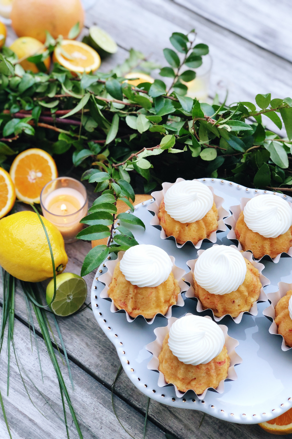 A citrus centerpiece with a white pedestal cake stand showcasing mini Bundt cakes topped with white frosting. Fresh greenery,  lemons, limes, and oranges are artfully arranged along the wooden table for an inviting and elegant display as table decoration.