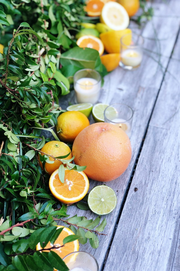 A vibrant citrus tablescape featuring fresh greenery, halved oranges, lemons, and limes, beautifully arranged along a rustic wooden table. Small votive candles add a warm glow to the citrus centerpiece ideal for a gender neutral baby shower idea.

