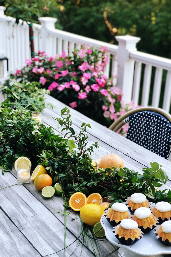 An outdoor citrus tablescape featuring a vibrant citrus centerpiece with fresh oranges, grapefruits, limes, and greenery arranged along the table. The natural wood table is surrounded by colorful flowers and lush greenery in the background. Soft candlelight complements the fresh citrus theme, creating a cozy yet elegant atmosphere for an orange baby shower or a summer citrus theme party.