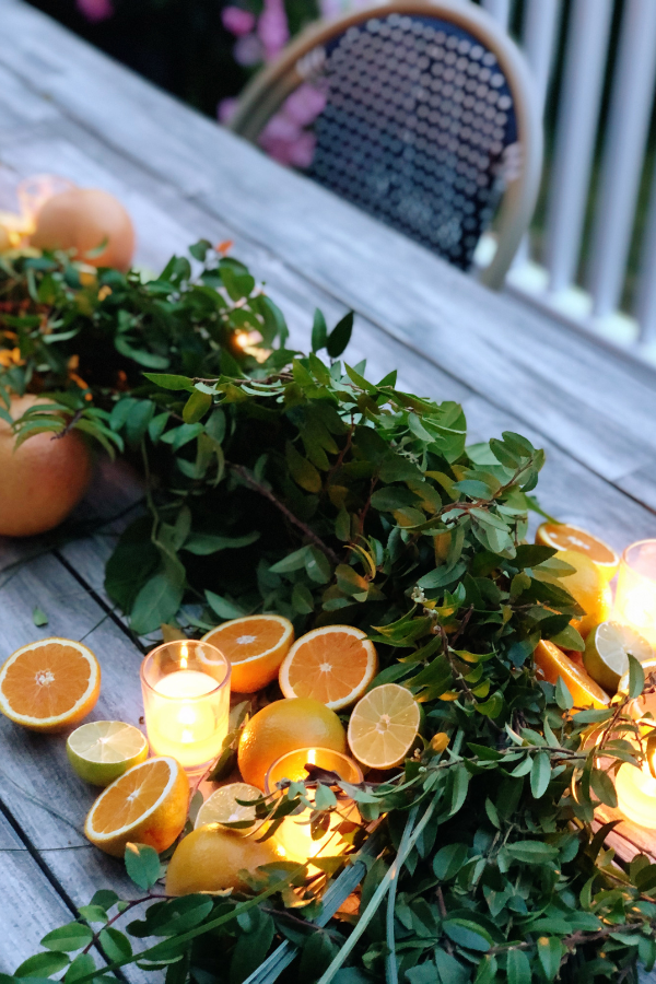 A citrus centerpiece with fresh greenery, sliced citrus fruits, and glowing votive candles are artfully arranged along the wooden table for an inviting and elegant display ideal for a summer citrus theme party.