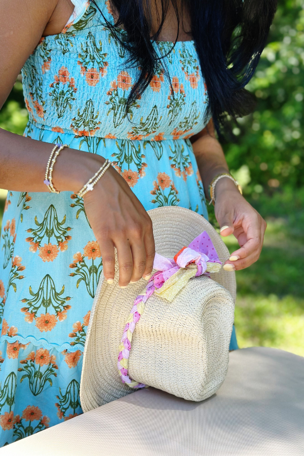 A woman in a blue floral dress is decorating a straw cowboy hat with braided fabric and a pastel bow. This coastal cowgirl aesthetic hat is perfect for cowgirl bachelorette parties, beach cowgirl hat ideas, and custom cowgirl hats.