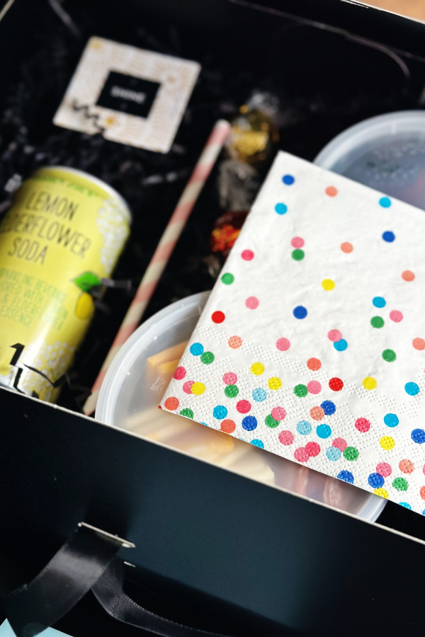 One black picnic box carefully packaged with charcuterie and fruit snacks along with a fortune, soda, straw and colorful polka dot napkin. 