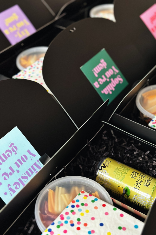 A group of cute black boxed lunches neatly arranged, each with colorful polka dot napkins, a can of lemon elderflower soda, and personalized stickers on the lids with each guests name on top. All tied together with a black ribbon.