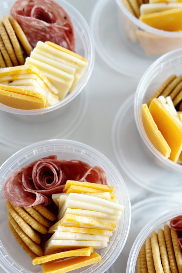 Clear plastic cups containing mini charcuterie boards, featuring slices of cheddar and colby jack cheese, crackers, and rolled salami arranged elegantly for single servings.