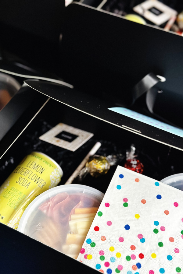 A collection of sleek black boxed lunches, each thoughtfully packed with a can of lemon elderflower soda, straws, chocolates, colorful polka dot napkins, charcuterie snack packs, and a “shine” fortune card. 