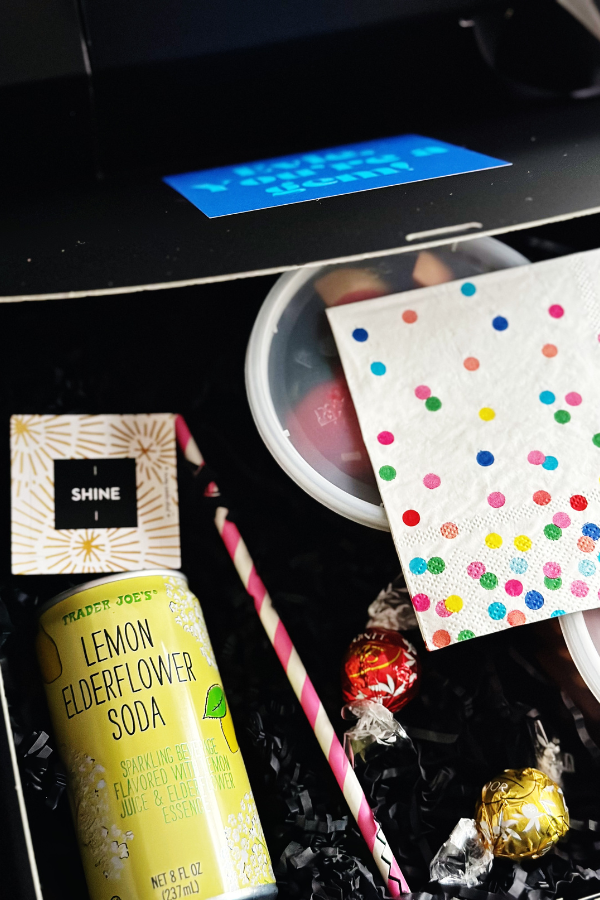 One black meal box carefully packaged with charcuterie and fruit snacks along with a "shine" fortune, soda, straw, chocolates, a colorful polka dot napkin and a personalized sticker on the top of the box.