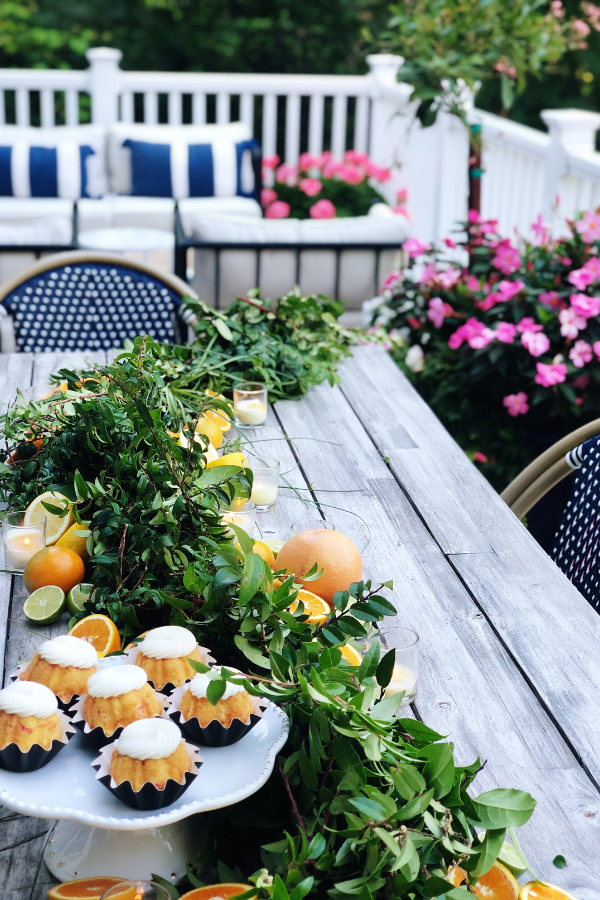 An outdoor citrus tablescape featuring a vibrant citrus centerpiece with fresh oranges, grapefruits, limes, and greenery arranged along the table. The natural wood table is surrounded by navy and white bistro chairs, with colorful flowers and lush greenery in the background. Soft candlelight complements the fresh citrus theme, creating a cozy yet elegant atmosphere for outdoor summer entertaining.