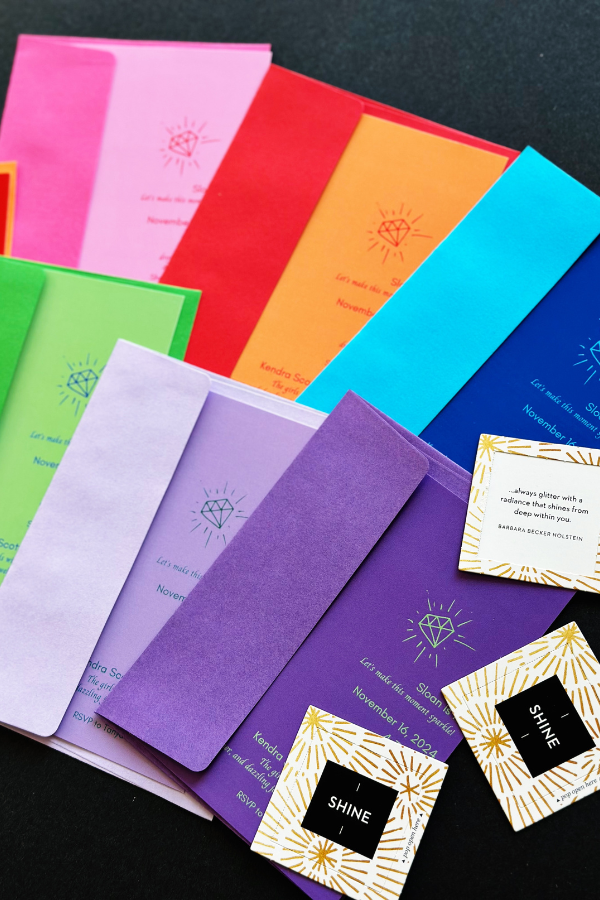 A flat lay of She’s a Gem birthday party invitations displayed on a black background. The cute invitations are in bold colors like orange, pink, purple, blue, and green featuring modern typography and a gem illustration. In the corner are small inspirational ‘Shine’ cards.