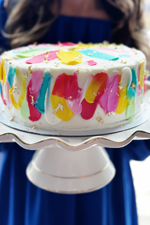 The photo features a jewelry party idea of taking a modern cake with many jeweled colors and applying edible gold leaf on top. The cake is sitting on top of a gold and white cake plate while a girl in a bright blue dress holds the cake.