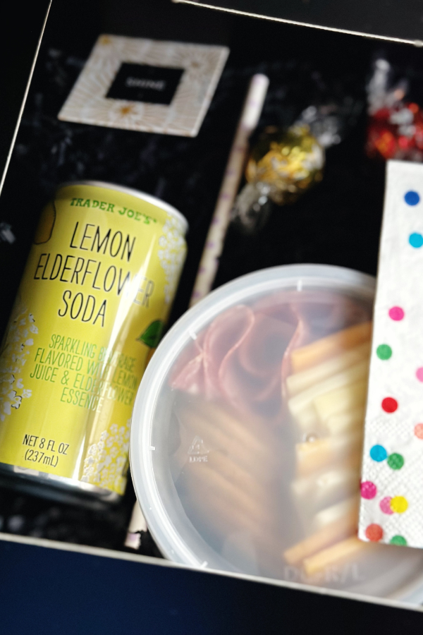 The photo featured a jewelry party idea of a cute black boxed lunch filled with crinkle paper, a yellow lemon elderflower soda, a paper straw, a container of cheese, crackers, and salami, a napkin with bright colored polka dots, chocolates, and a fortune tucked inside the box.