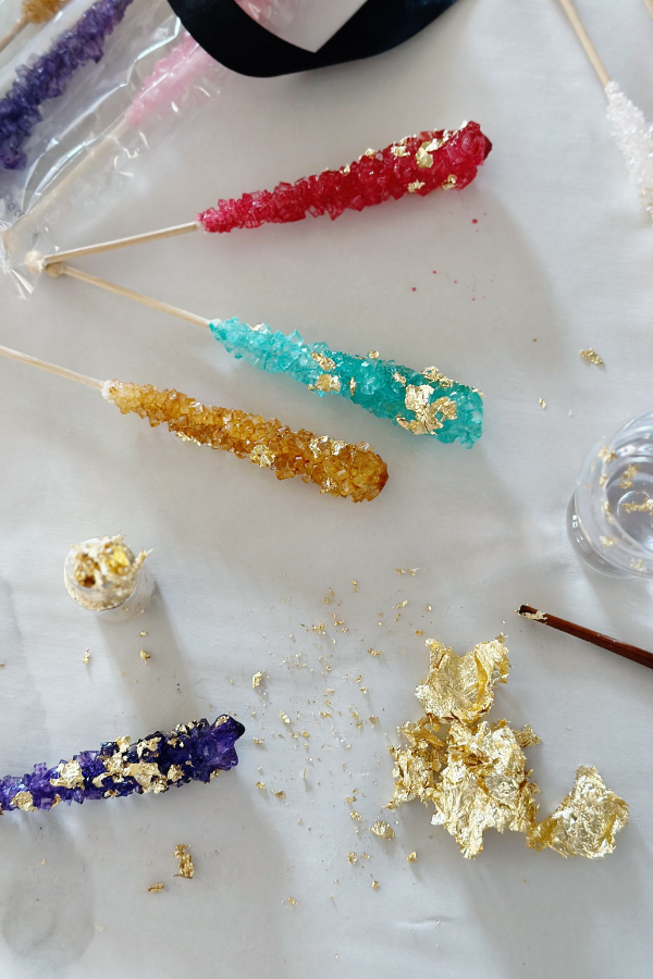 The photo features a jewelry party idea of taking rock candy sticks in red, blue, gold and purple and applying edible gold leaf to the candy treats.