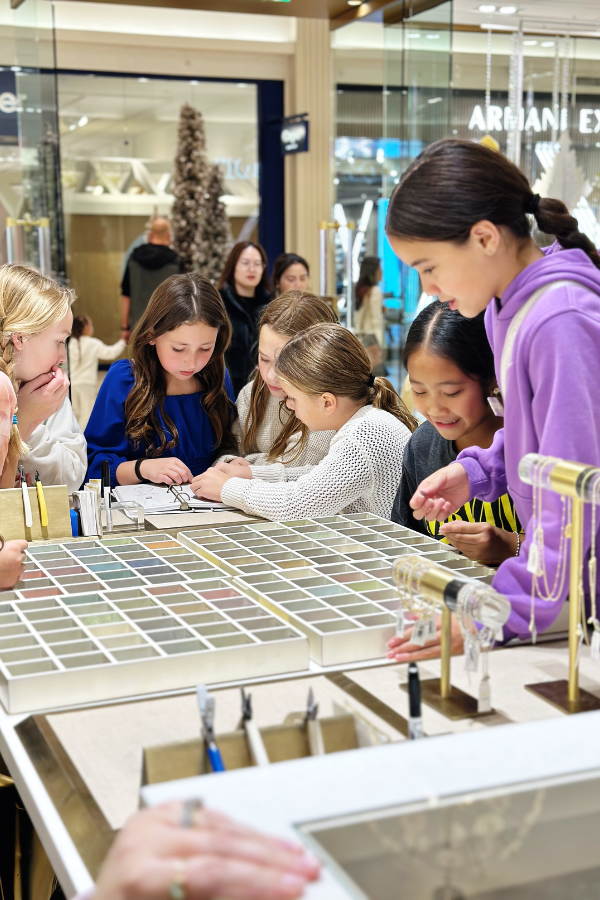 The photo features eleven year old girls at a jewelry making party at the Kendra Scott Color Bar. The girls are surrounding a DIY jewelry display looking at several colorful gem stones.