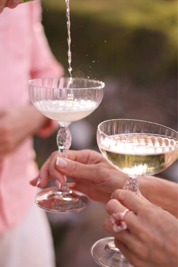 A close-up of two hands holding elegant coupe glasses filled with champagne during a bridal shower celebration. One glass is being filled with bubbly, capturing the light as it sparkles. The hands have manicured nails, and one features a pink floral ring, adding a romantic touch. The blurred background suggests an outdoor gathering with guests dressed in soft, festive tones.