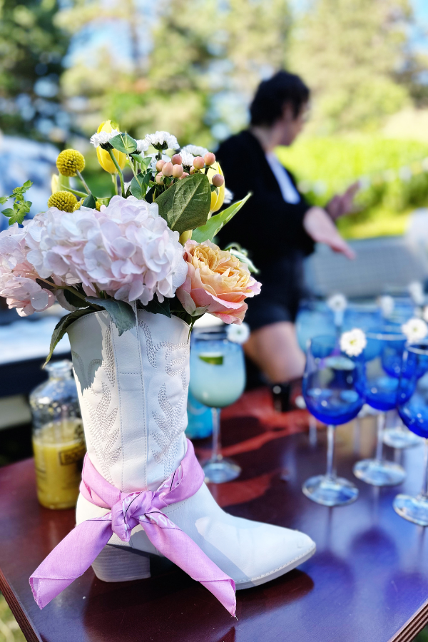 Cowgirl bridal shower decor featuring a flower arrangement set in a white cowboy boot with a pink bandana tied around it. This boot with flowers centerpiece adds a rustic touch to the drink station, perfectly complementing the western bridal shower ideas. 