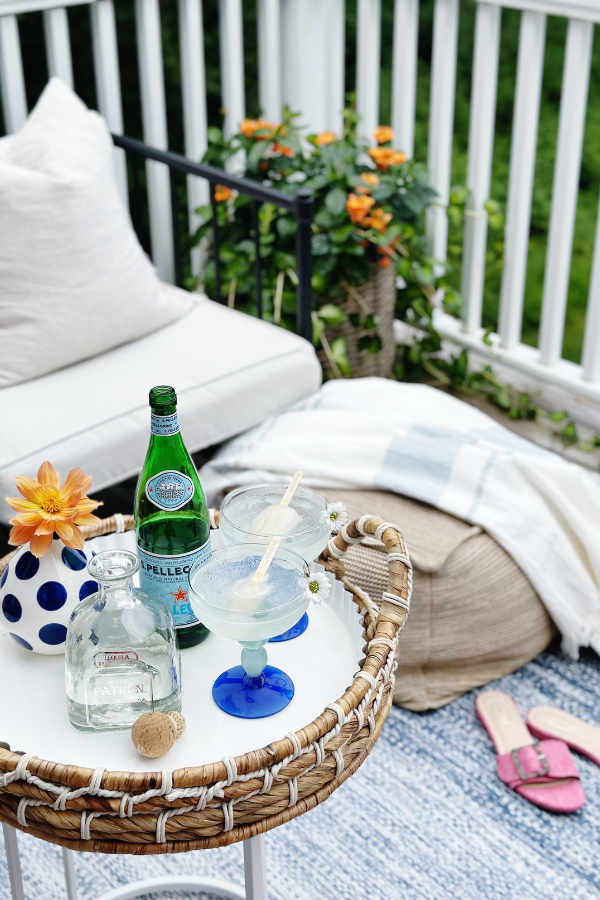 A bright and airy outdoor scene featuring a cozy patio setup with a textured rug, neutral throw pillows, and a woven tray displaying Texas Ranch Water cocktails with lime popsicles, tequila, and a bottle of sparkling water. Pink sandals rest nearby, adding a casual summer vibe.