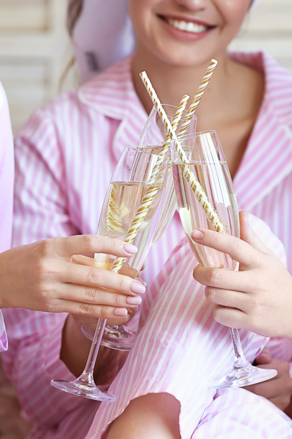 The photo is of a bride to be dressed in pink and white stripe pajamas toasting with her girlfriends at a wedding shower where they are enjoying a bridal shower brunch.