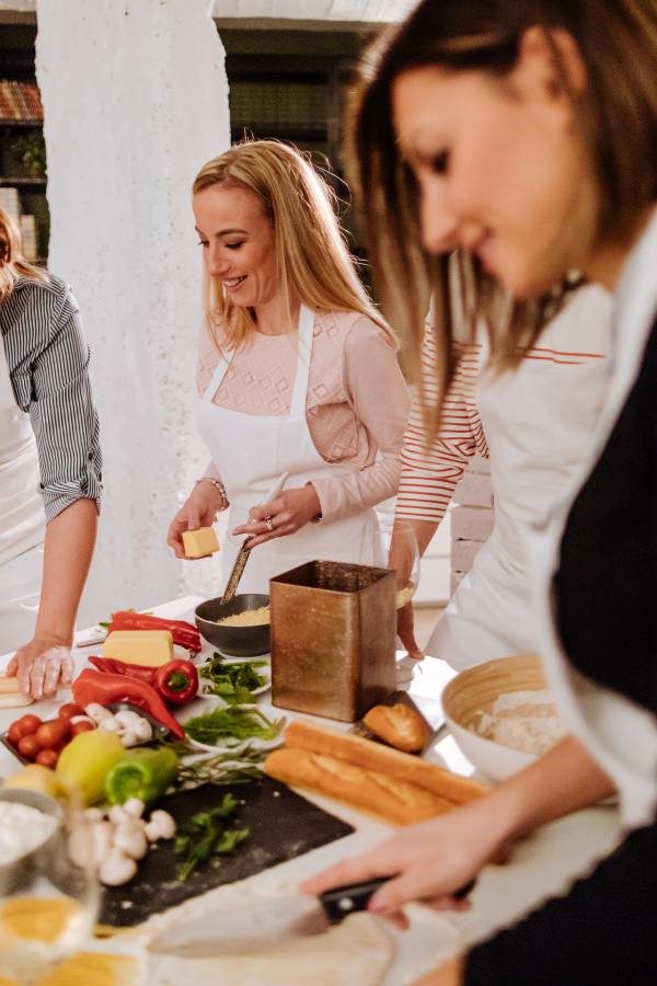 Instead of participating in wedding shower games guests at this bridal shower are in a private cooking class. One guest is rolling out dough and one is grating cheese while others look on.