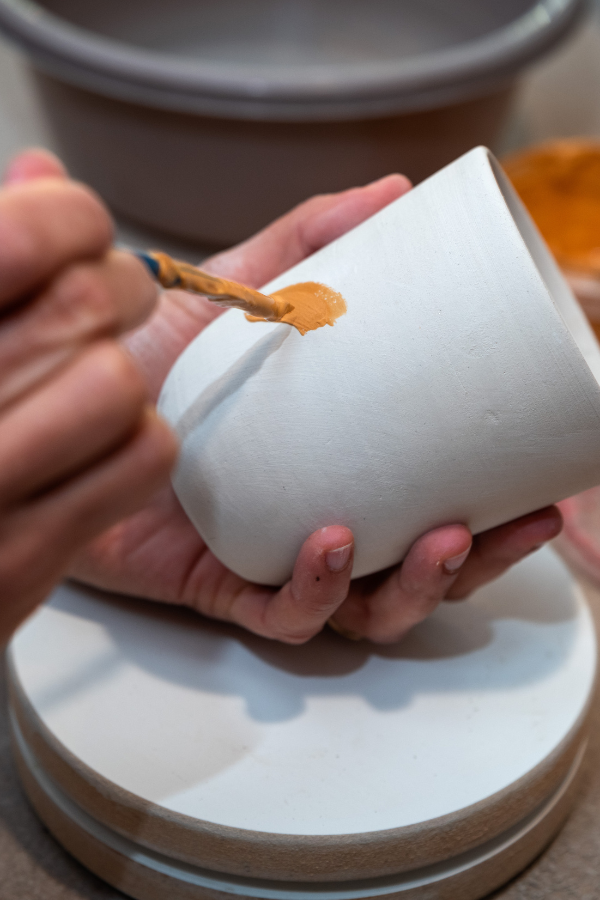 The photo is close up of a women's hands holding a piece of pottery in one hand and a paint brush with mustard yellow paint in the other. There is one paint stroke on the piece of pottery.