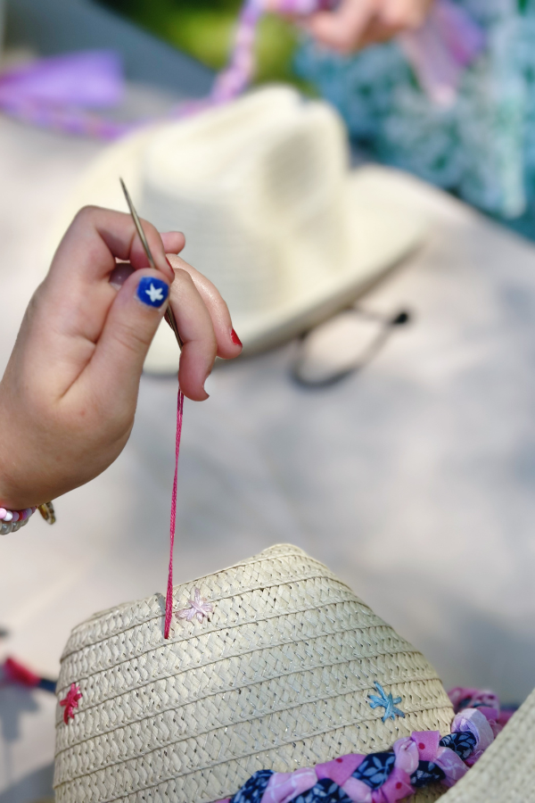 A close-up of a child’s hand sewing colorful embroidered stars onto a beachy cowboy hat with braided fabric trim. 