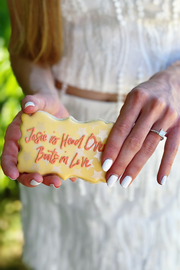 Close-up of the bride-to-be at her wedding shower holding a cut-out cookie that reads "Josie is Head Over Boots in Love." 