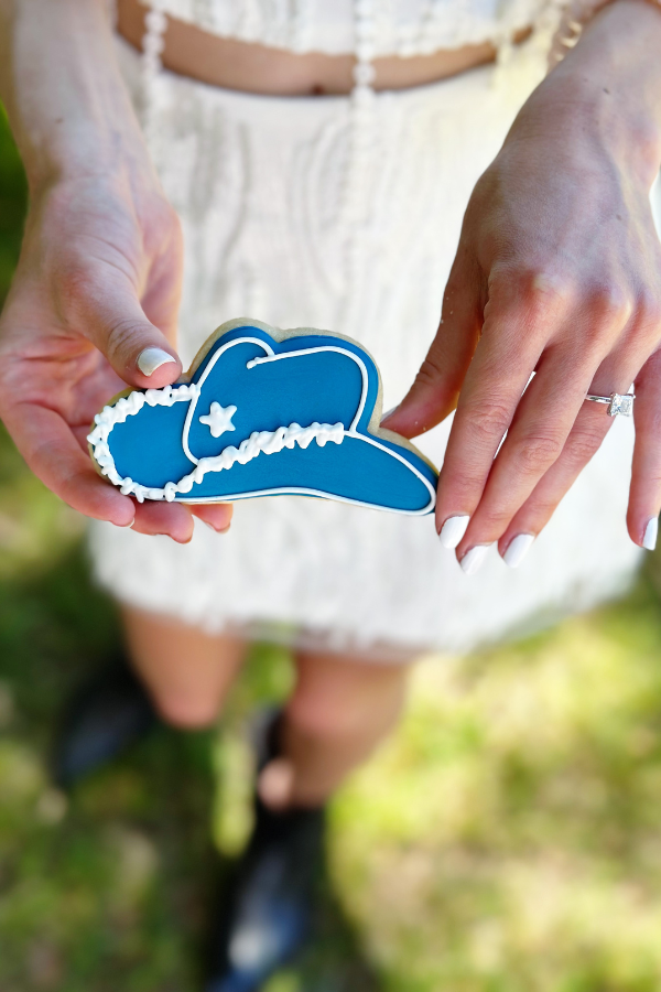 Close-up of the bride-to-be holding a frosted blue cookie shaped like a cowgirl hat at her cowgirl bridal shower. The cookie, decorated with intricate details is perfect for the cowboy theme party.