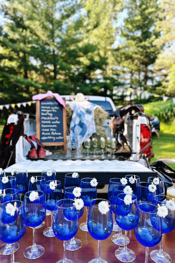 A rustic outdoor cowgirl bridal shower drink station featuring cobalt blue wine glasses with white daisy accents. A pickup truck bed serves as a bar, decorated with hay bales, cowboy boots, and a saddle, perfect for Western bridal shower ideas or a cowgirl-themed bachelorette party. A handwritten chalkboard menu adds a charming boho bridal shower touch.