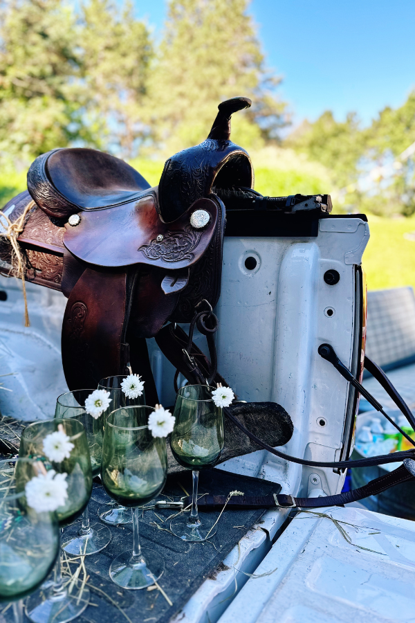 A rustic outdoor cowgirl bridal shower drink station featuring green wine glasses with white daisy accents. A white pickup truck bed serves as a bar, decorated with and a saddle and a hay bail.