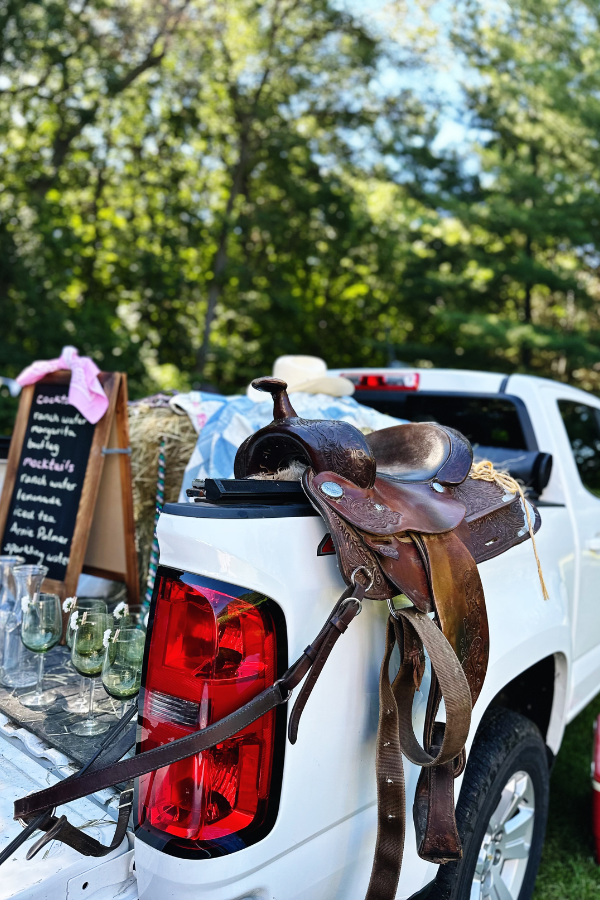 A rustic outdoor cowgirl bridal shower drink station featuring a leather saddle on top of a pickup truck. The bed of the truck serves as a bar, decorated with hay bales and a denim quilt, perfect for Western bridal shower ideas or a cowgirl-themed bachelorette party. A handwritten chalkboard menu adds a charming boho bridal shower touch.