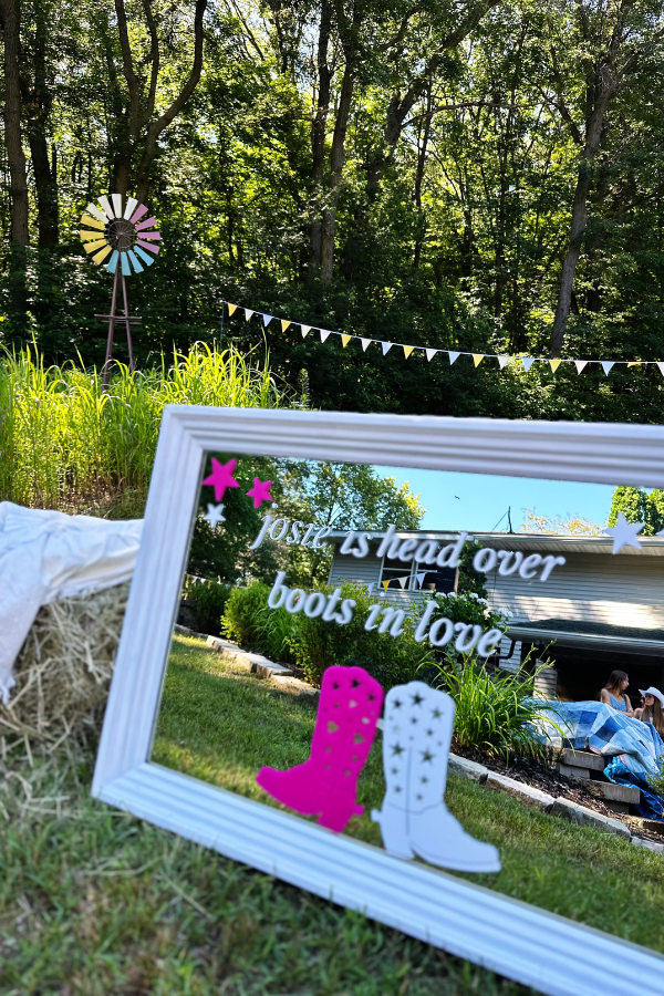 Rustic decorations including a windmill, yellow pendant flags, and a mirror with the phrase 'Josie is Head Over Boots in Love' propped against a hay bale. This beautiful backyard scene features boots and bubbly bridal shower ideas and western bridal shower ideas, ideal for a cowgirl-themed bachelorette party or rustic bridal shower decorations. 