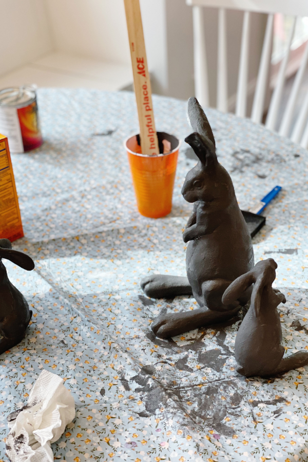 A DIY Easter craft decorations project in progress, featuring several Easter bunny crafts painted in a dark charcoal gray. The table is covered with a floral-patterned tablecloth, and supplies like paintbrushes, orange Solo cups, a box of baking soda, and a wooden stir stick are scattered around. These homemade Easter decorations are an easy Easter craft idea, perfect for adding a modern touch to Easter home decor.