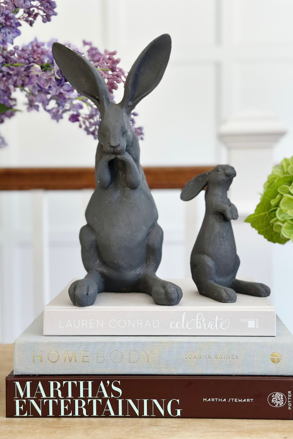 Close-up of two DIY Easter bunny crafts sitting atop a stack of three home decor books, styled as part of elegant Easter home decor. The bunnies have a modern, textured finish created using an easy Easter decorations DIY technique with paint and baking soda. In the background, a green hydrangea and a purple lilac add a touch of spring decor. This homemade Easter decoration is a perfect example of cute Easter crafts and Easter craft decorations that elevate your space.