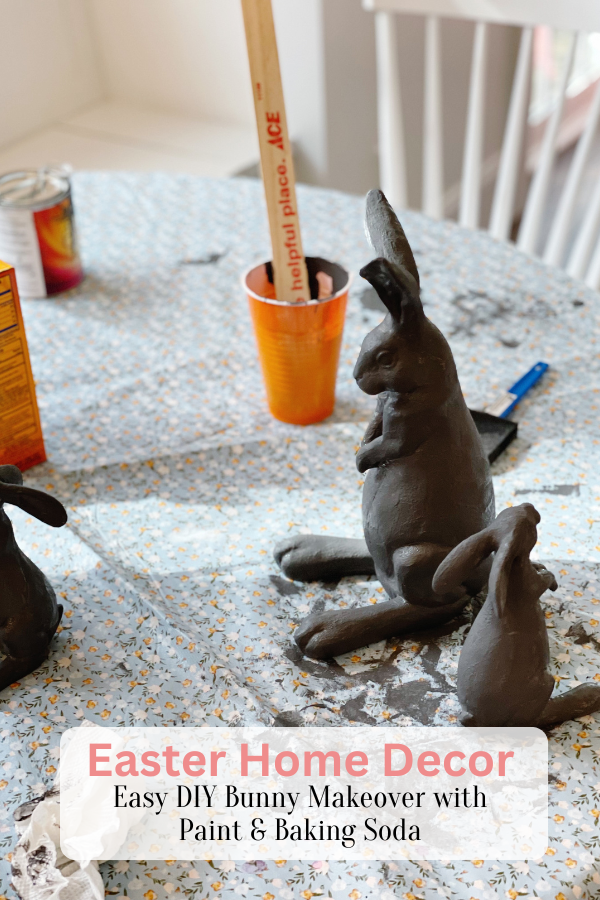 A DIY Easter craft decorations project in progress, featuring several Easter bunny crafts painted in dark gray. The table is covered with a mini floral-patterned tablecloth, and supplies like paintbrushes, orange cups, a box of baking soda, and a wooden stir stick are scattered around. The text overlay reads "Easter Home Decor | Easy DIY Bunny Makeover with Paint & Baking Soda".