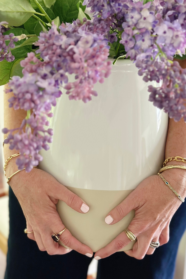 A close up picture of a woman with gorgeous gold jewelry and perfectly manicured nails holding a large stoneware vase overflowing with lavender lilac stems showcasing modern Easter decor that's both neutral and organic for spring decoration. 