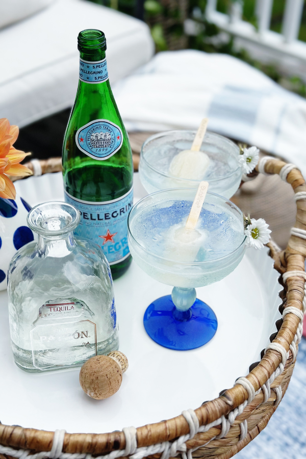 A cozy outdoor summer setup with a white tray holding sparkling water cocktails, tequila, and a little daisy garnish. A soft striped throw and woven pouf add texture, making it the perfect setting for weekend therapy with a refreshing cocktail.