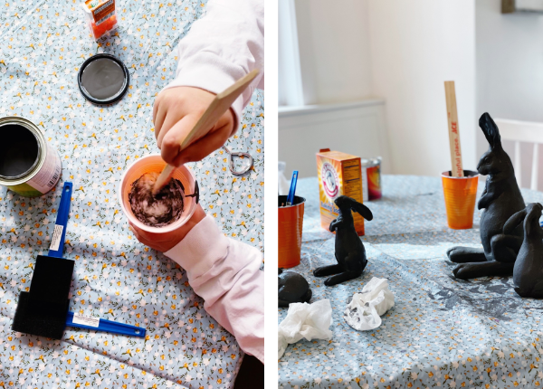 The image is split into two on the right is a DIY Easter craft decorations project in progress featuring an open paint can and a few foam paint brushes while a little girl mixes paint and baking soda together in an orange Solo cup. The right side of the image features several Easter bunny crafts painted in dark gray. The table is covered with a floral-patterned tablecloth, and supplies like paintbrushes, orange cups, a box of baking soda, and a wooden stir stick are scattered around. These homemade Easter decorations are easy Easter crafts for kids and a perfect modern touch to Easter home decor.