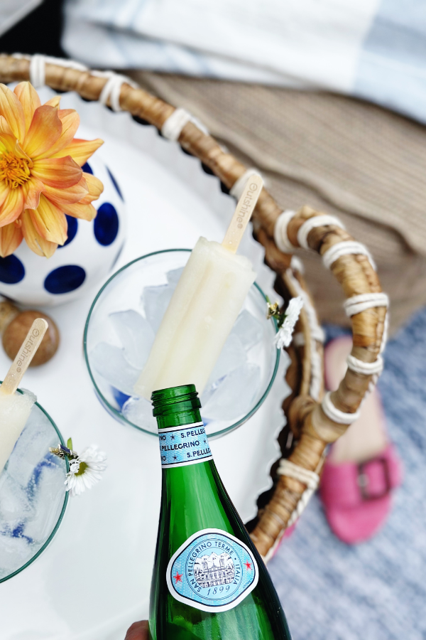 A close-up of a ranch water popsicle cocktail with an Outshine lime popsicle resting in a glass filled with ice and sparkling water. A hand pours from a green bottle of San Pellegrino, adding the perfect fizzy touch to this light cocktail recipe.