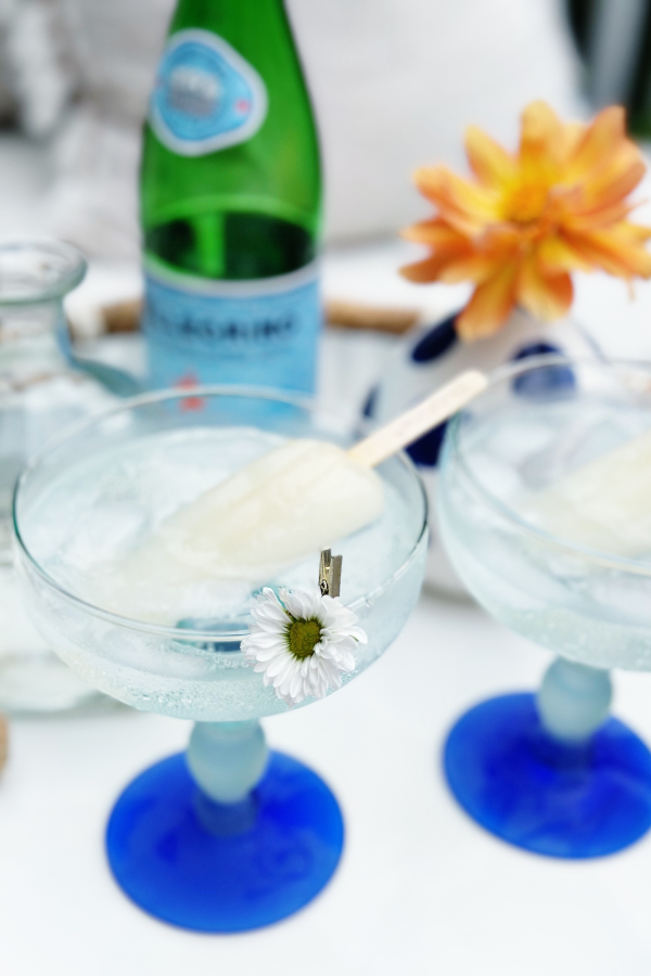 A close-up of two clean cocktails served in icy margarita glasses, each with a lime margarita recipe twist using lime popsicles and sparkling water. A green bottle of sparkling water along with an open bottle of tequila creating a refreshing, healthy popsicle recipe.