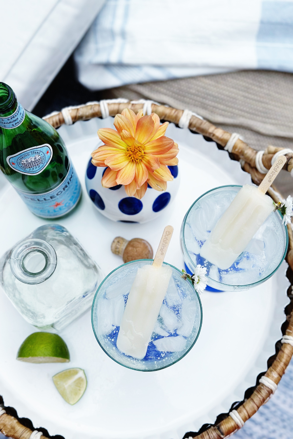 A woven rattan tray holding two ranch water popsicle cocktails served in blue-rimmed margarita glasses, each with a pale yellow lime popsicle melting into sparkling summer drink. A bottle of tequila, a green bottle of sparkling water, and a small white vase with orange flowers add a summery touch.