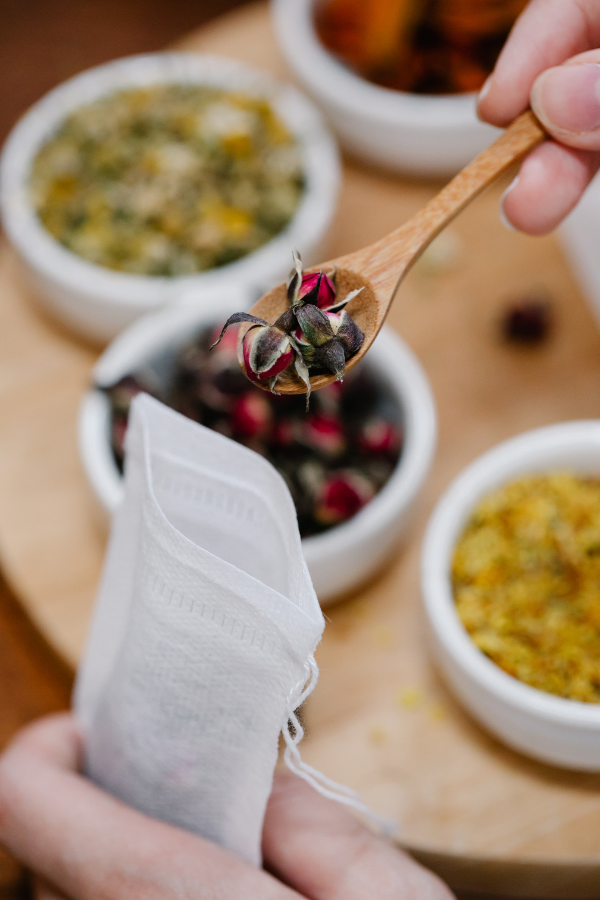 A tray of dried flower and herbs is displayed in the background and a small wooden spoon is putting dried rose buds into a sachet as an example of a small bridal shower idea.