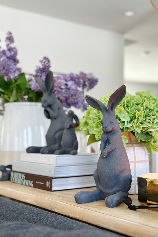 Styled console table featuring four DIY Easter bunny crafts as part of elegant Easter home decor. The handmade bunnies, created using an easy Easter decorations DIY technique with baking soda and paint, add a modern touch to the display. A vase with lilacs and hydrangeas brings a fresh spring element, complementing the neutral tones of the bunnies. 