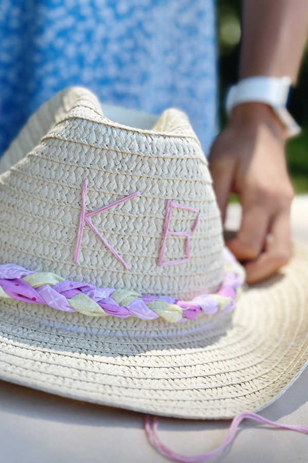 A close-up of a beautifully decorated straw cowboy hat, featuring hand-stitched initials KB and a braided fabric trim in pinks and yellows. This embellished cowboy hat project is perfect for a cowgirl birthday or a cowgirl bachelorette party. The design blends a vintage cowgirl vibe with a straw hat aesthetic, making it an eye-catching accessory for a beach cowgirl hat look or a last toast on the coast celebration.