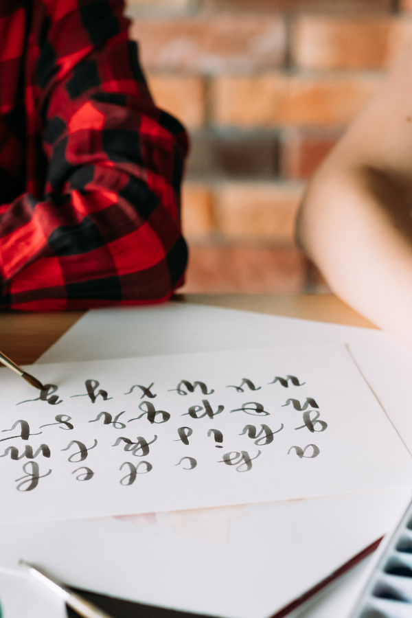 A women in a buffalo plaid shirt is learning calligraphy. Her beautiful hand lettering is an example of a wedding shower activity verses playing bride shower games.