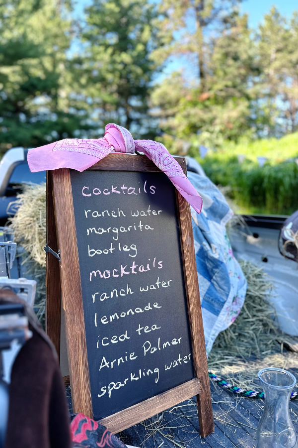 Chalkboard sandwich board sign at a  drink station featuring signature drinks for a cowgirl bridal shower. The menu includes creative beverages for a western bridal shower, adding a rustic touch. 