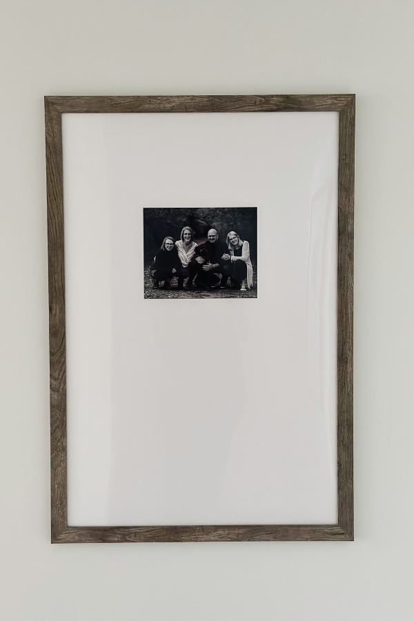 A large picture frame with a neutral wood border, showcasing a black-and-white family portrait with an oversized white mat in the frame. A simple and elegant wall decor idea for a small hallway or gallery wall inspiration. The picture is offset from the center giving the artwork a high-end look. 