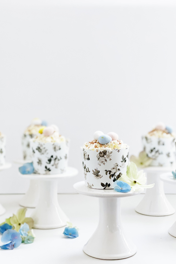 A pastel-hued spring cupcake sits atop a white pedestal, nestled in a chic black floral wrapper. The cupcake’s smooth white buttercream is topped with chocolate shavings. Soft green and blue florals are scattered throughout the photo reinforcing a stunning Easter-themed dessert for a sophisticated gathering.