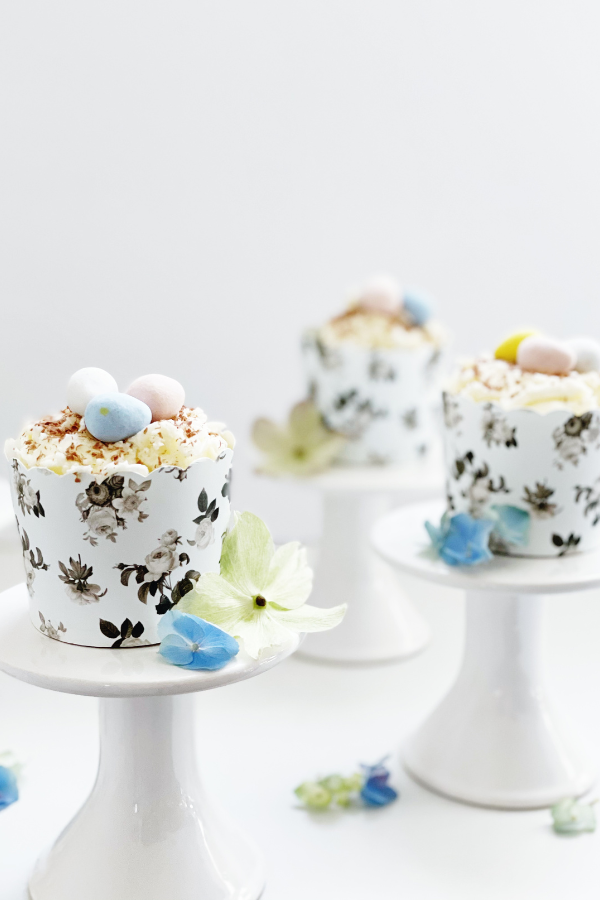 An elegant Easter cupcake idea featuring a white-frosted cupcake with chocolate shavings, resting in a black floral wrapper with blue and green florals scattered about. The cupcake is placed on a crisp white pedestal, this exquisite treat embodies the beauty of the season, making it a must-have spring dessert for Easter brunch.